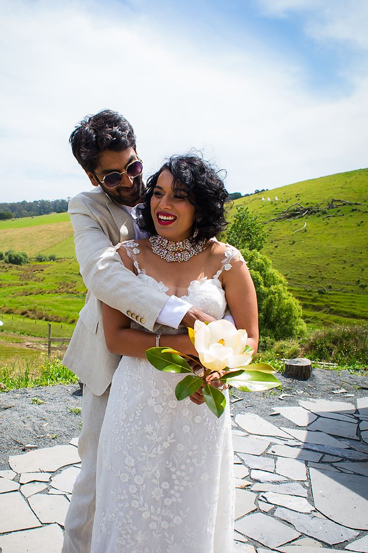 Newlyweds share joyful laughter during outdoor photoshoot with rolling green hills and blue sky in background.