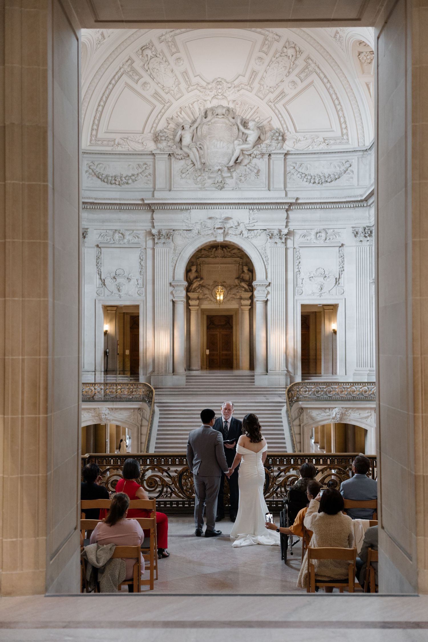 Elegant wedding ceremony taking place in a grand marble staircase hall with ornate architectural details and white walls.