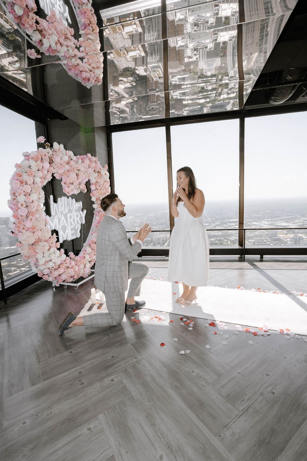 A marriage proposal moment with pink floral decorations in a modern high-rise space overlooking the city.