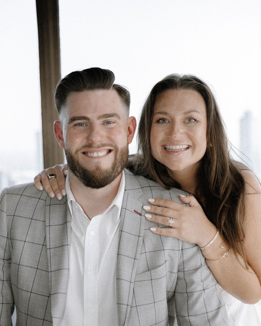 Happy couple smiling and showing off engagement ring in casual indoor setting.