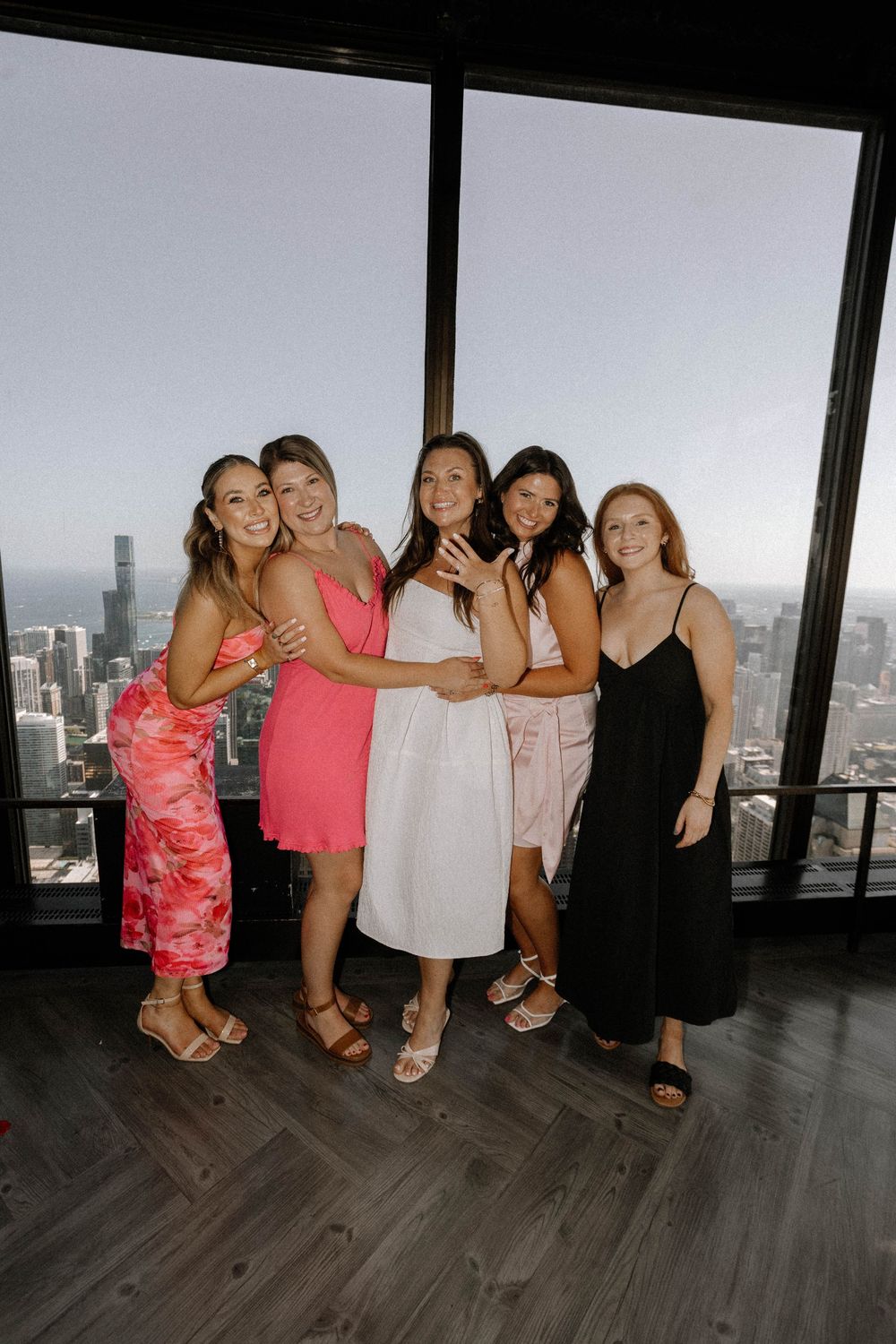 A group in summery attire poses playfully on a wooden floor with a city skyline view through large windows behind them.