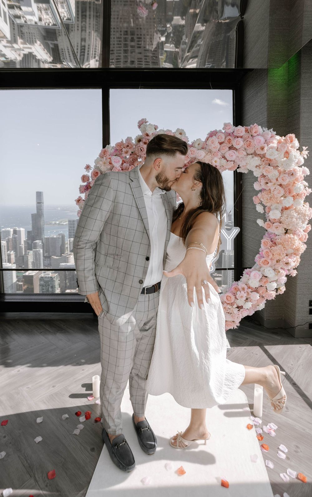 Couple poses romantically by pink floral arch in modern high-rise setting with city views.