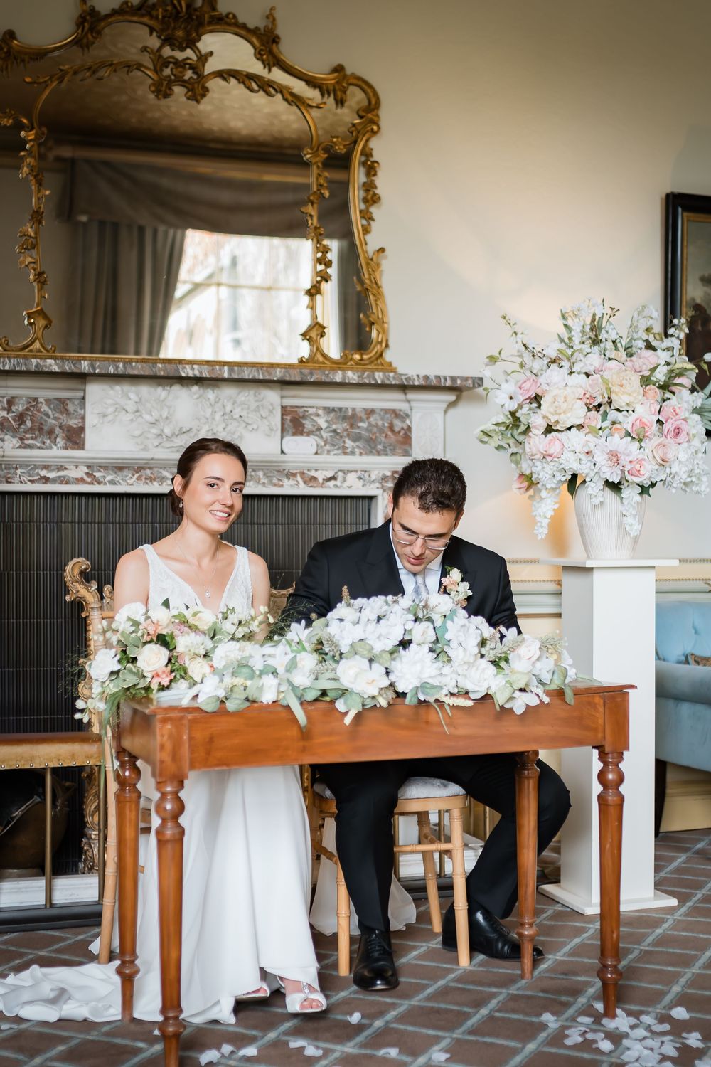 Elegant wedding venue decorated with ornate gold mirrors and white floral arrangements on wooden tables.
