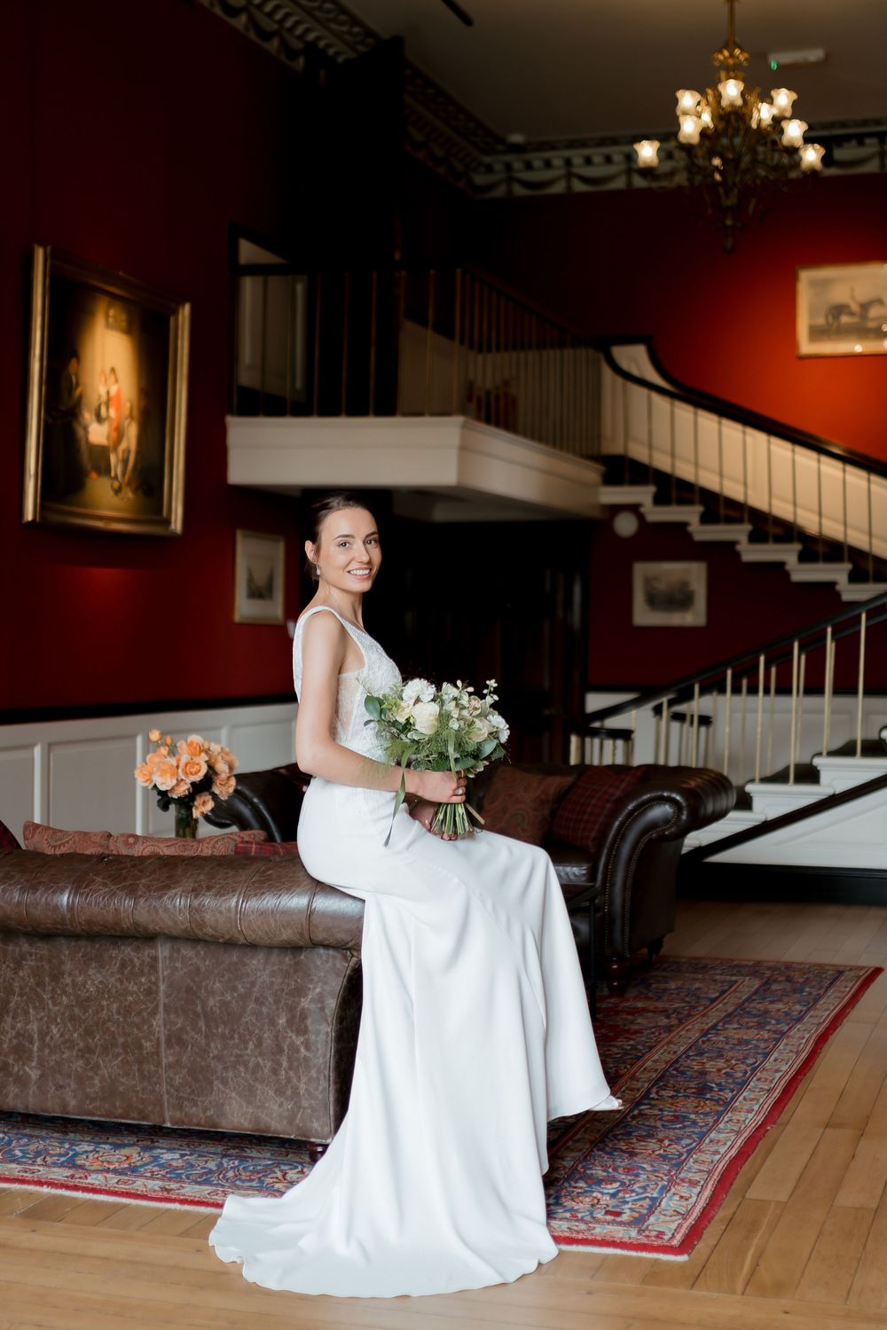 A bride in a white wedding gown sits gracefully on a vintage couch in an elegant room with red walls and hardwood floors.