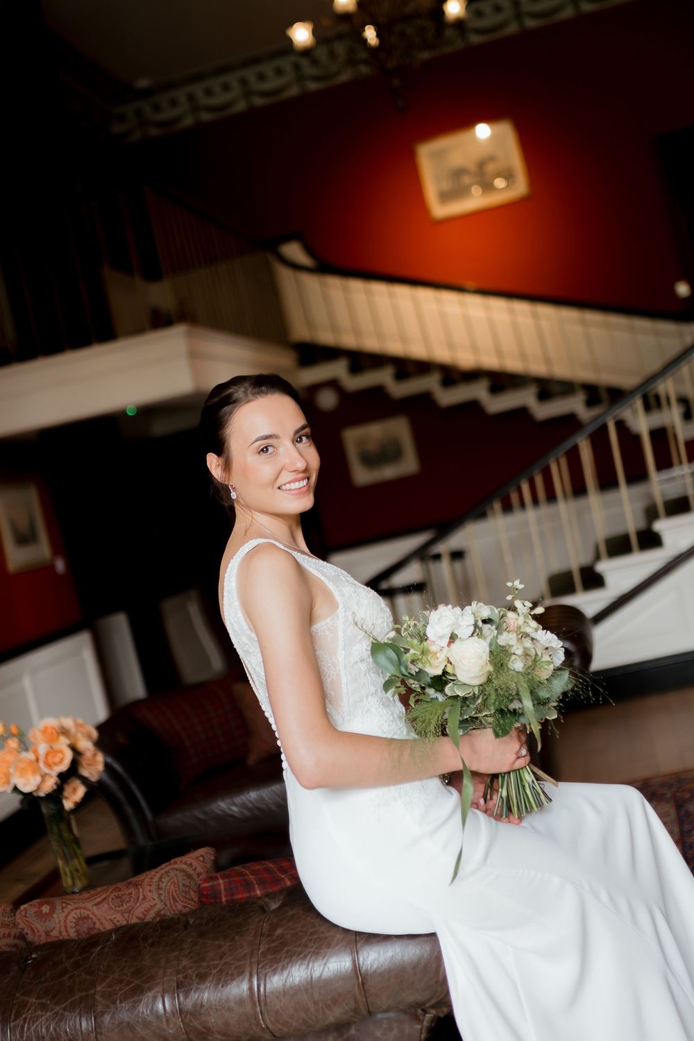 A bride in an elegant white dress sits on a brown leather couch holding a beautiful bouquet of white and green flowers.