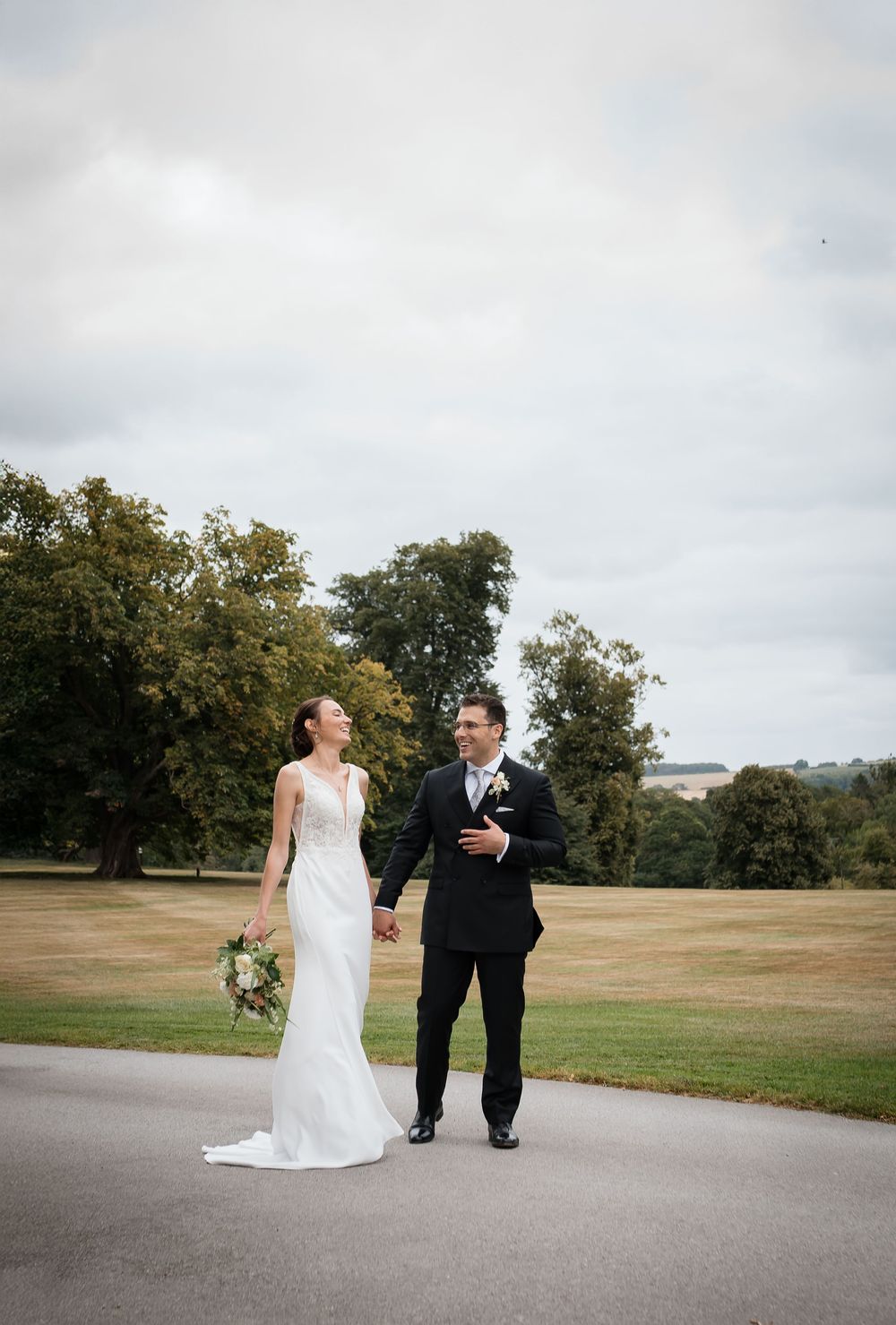 A couple in wedding attire walk and laugh together on a paved path surrounded by green lawns and trees.