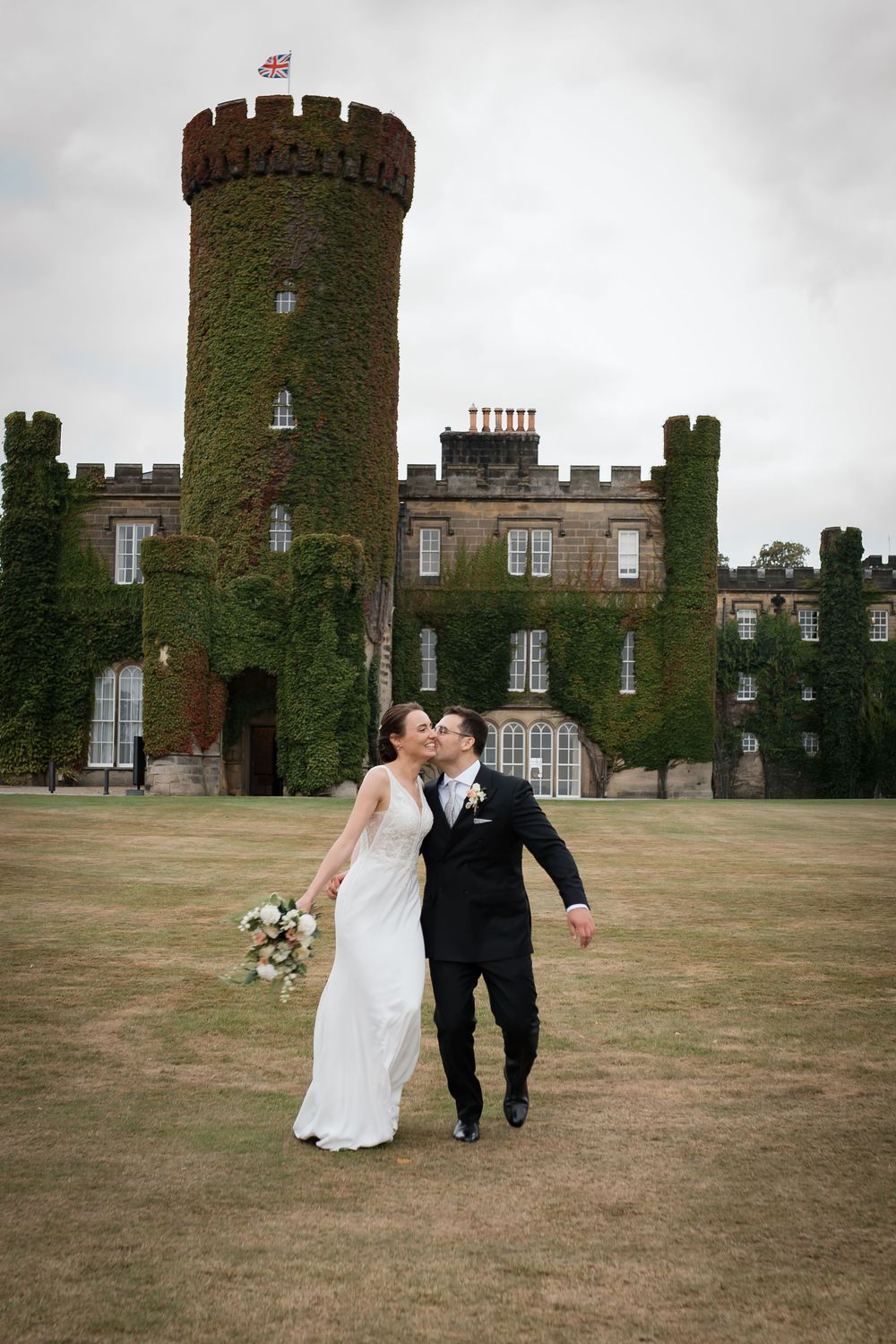 A couple walks hand in hand across the lawn of a majestic stone castle with ivy-covered towers and crenellated walls.