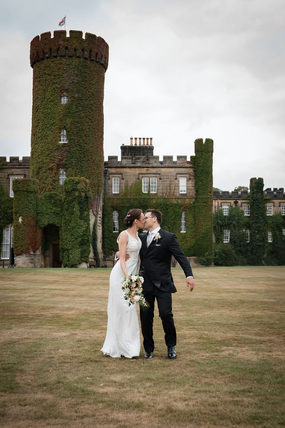 A newlywed couple shares an intimate moment in front of an ivy-covered castle with towers and turrets.