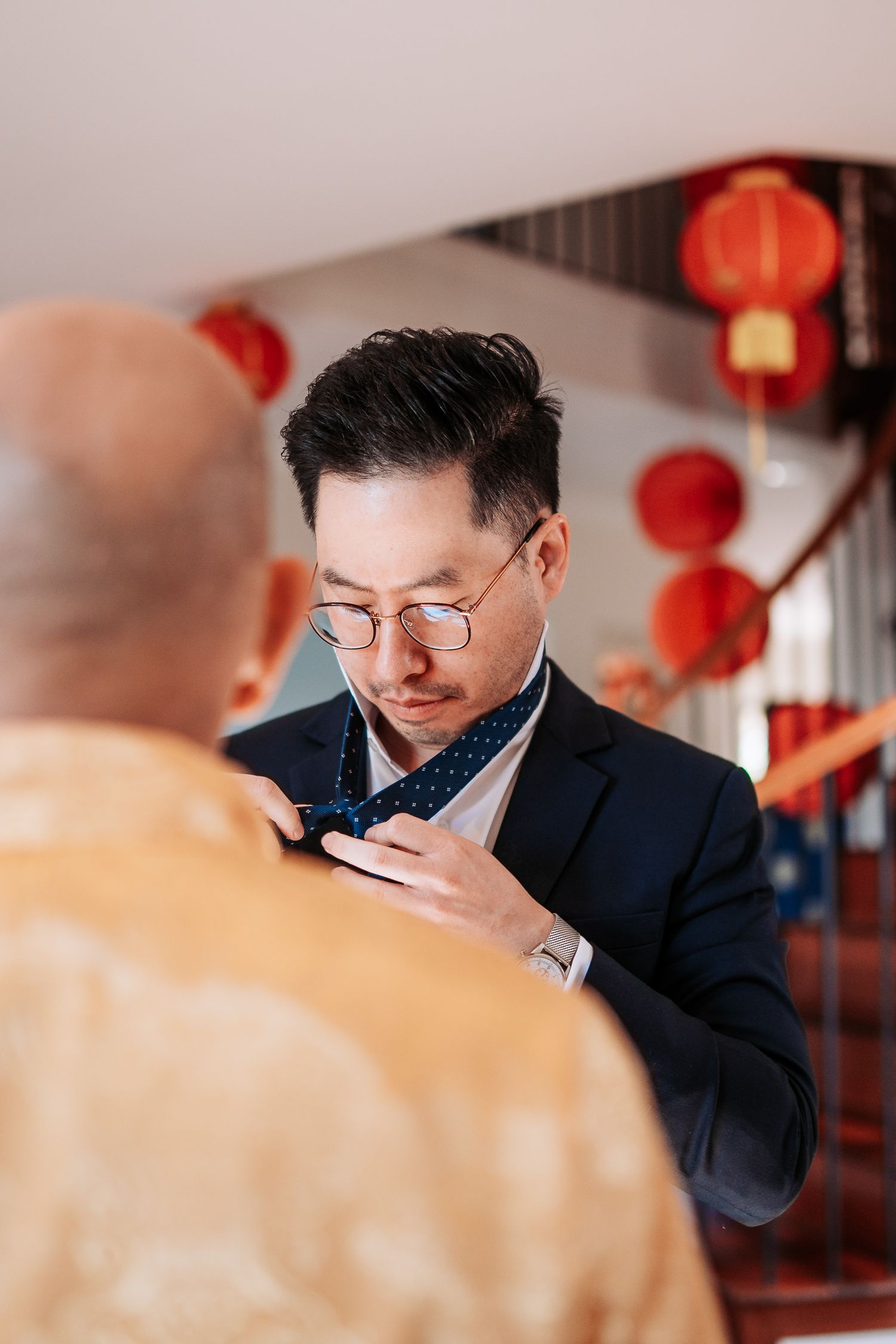 Man in suit adjusts formal attire with red lanterns decorating the background.