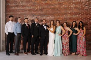 Wedding party members stand in a row against a rustic brick wall backdrop in formal attire.
