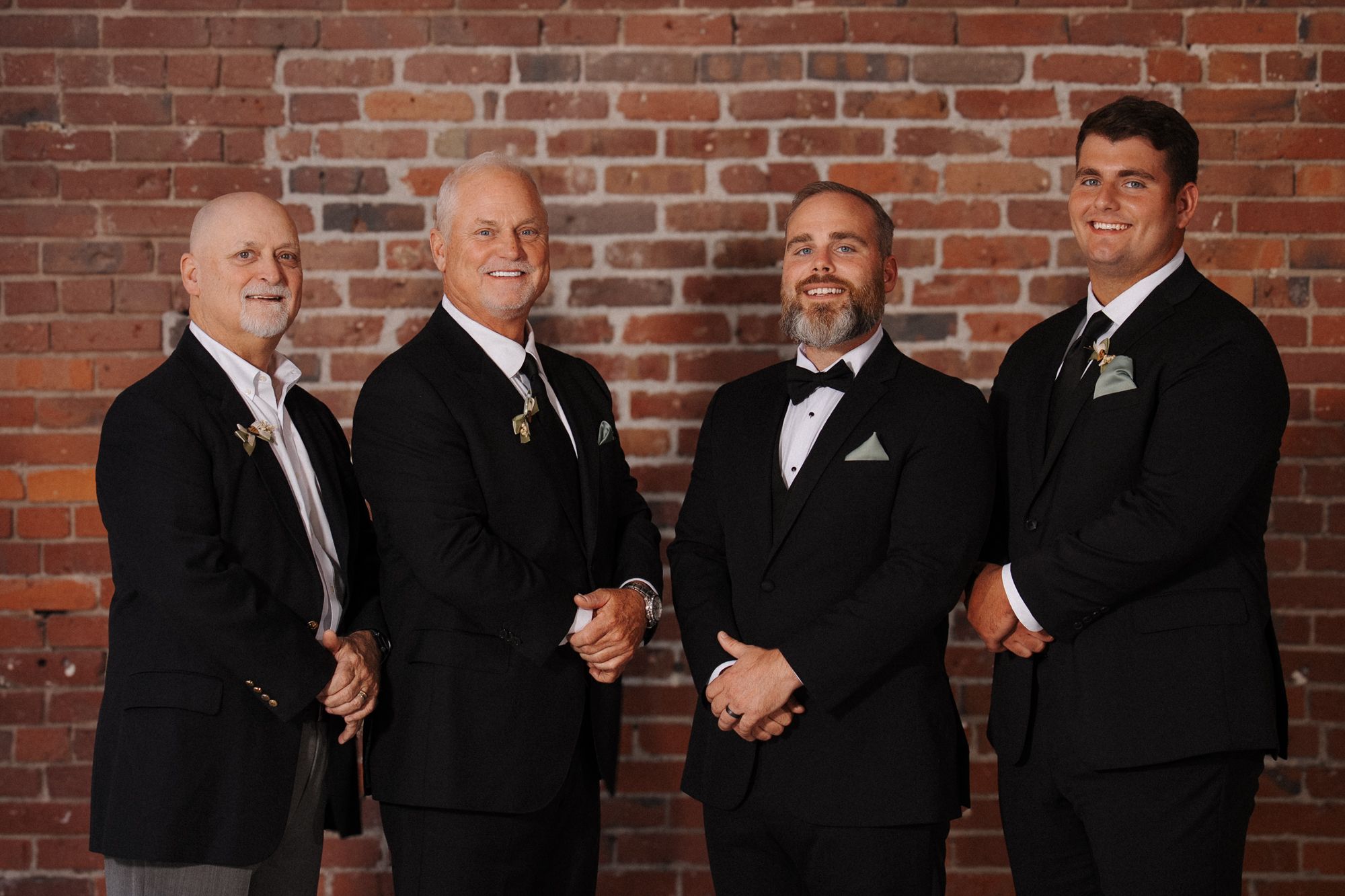 Wedding party wearing black tuxedos poses in front of red brick wall background.