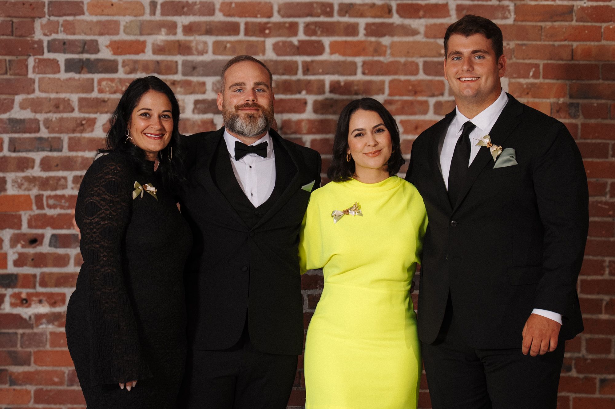 Wedding party group photo against a rustic brick wall with formal attire and bright yellow accent.