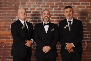 Three men in formal black suits and bowties stand together against a rustic brick wall backdrop at an event.
