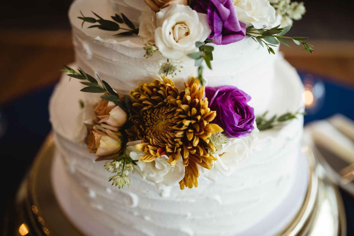 White wedding cake decorated with fresh purple and white flowers and gold accents on display at reception.