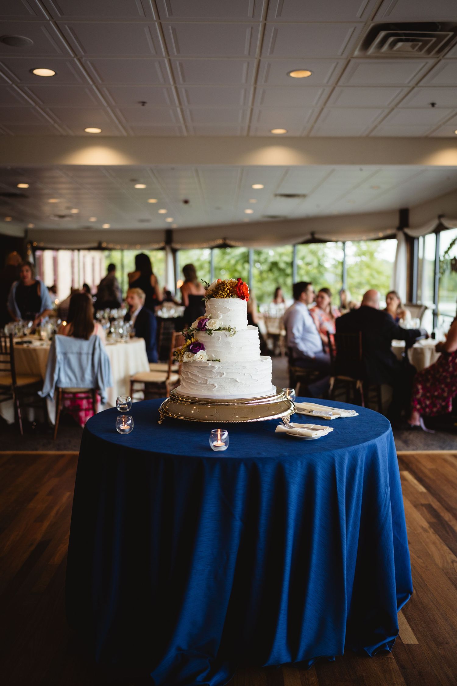 White wedding cake displayed on blue tablecloth in wedding reception venue with warm lighting and wooden floors.