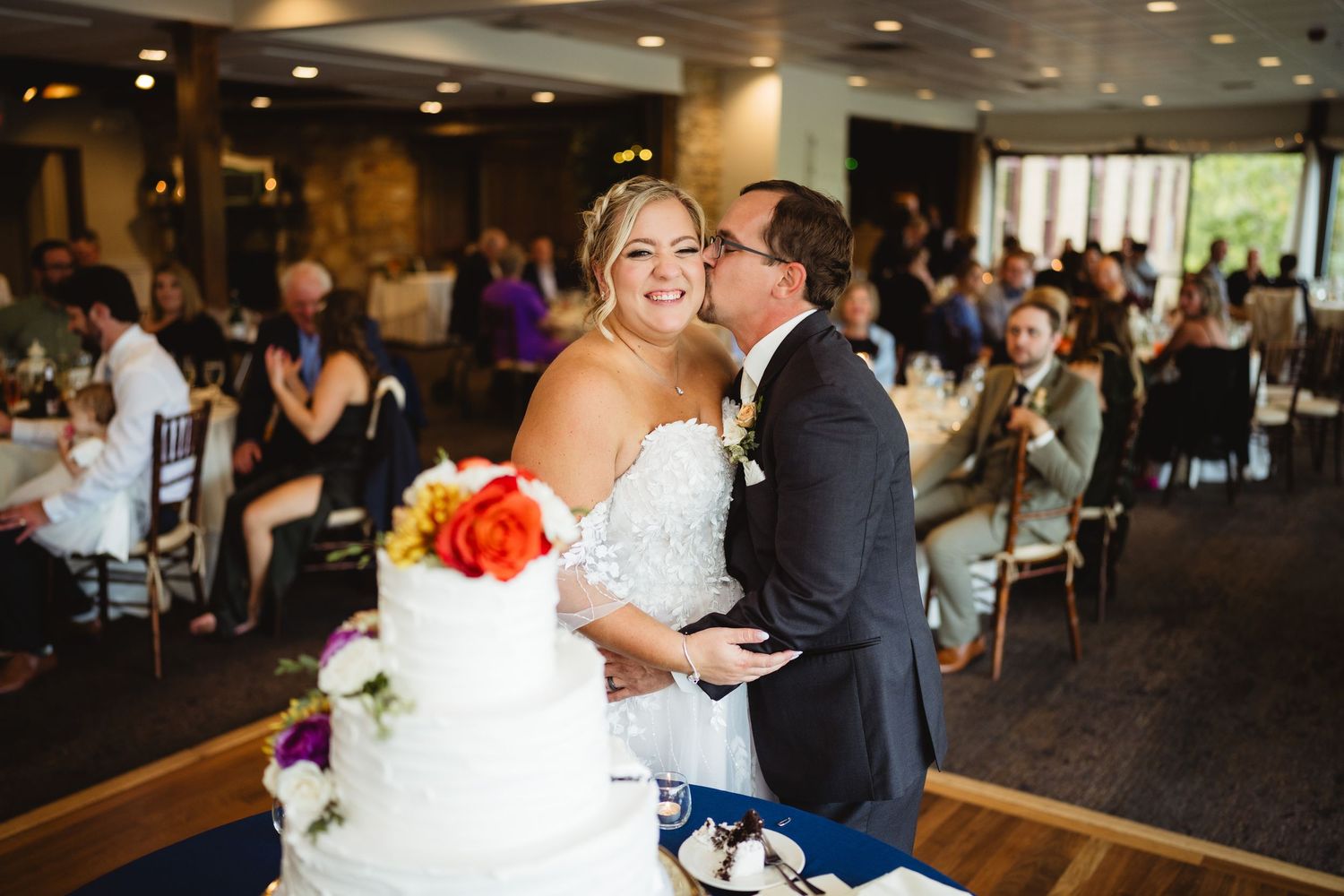 A couple shares a tender moment at their wedding reception by the cake decorated with orange flowers in an elegant venue.