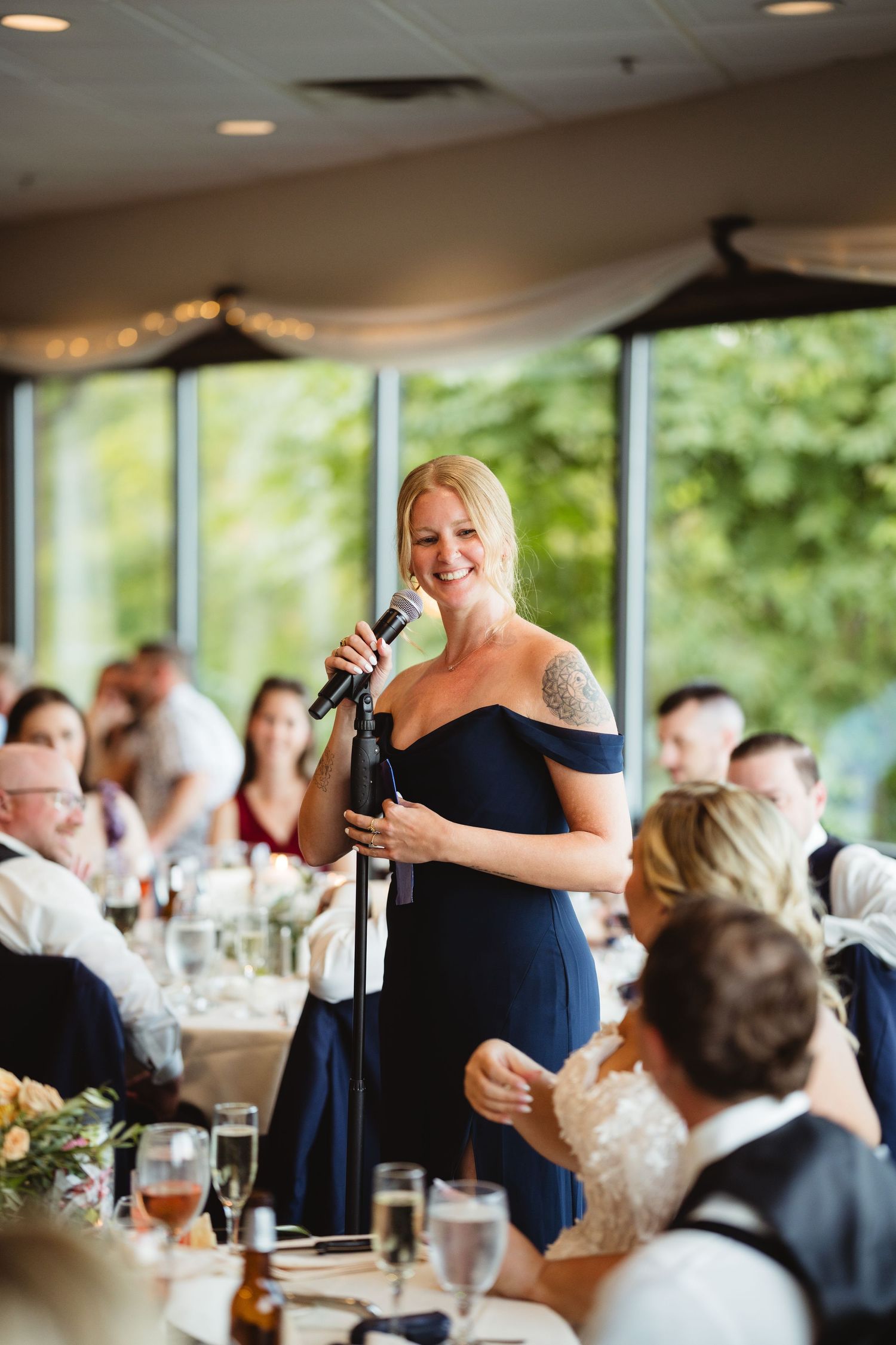 Bridesmaid in navy dress giving emotional wedding speech at reception dinner while guests listen attentively.