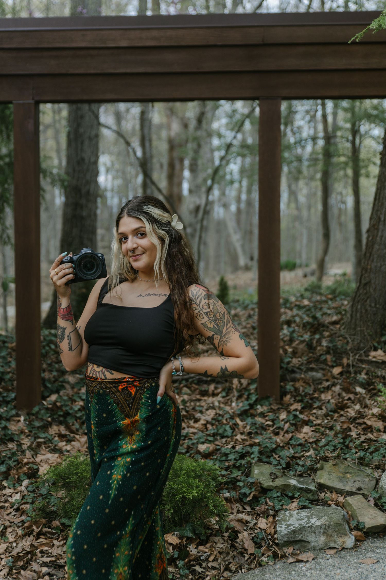 Woman in boho style clothing holding a camera in a forest setting with wooden beams overhead.