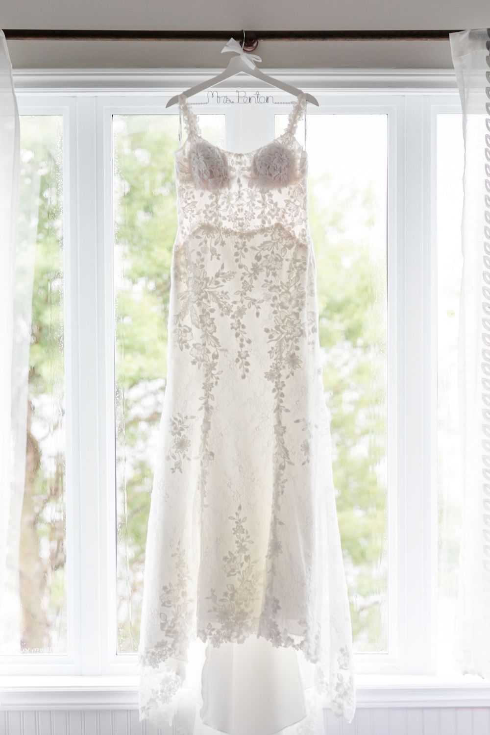 Several elegant white wedding dresses with lace and embellishments hang in a bright, sunlit room by windows.