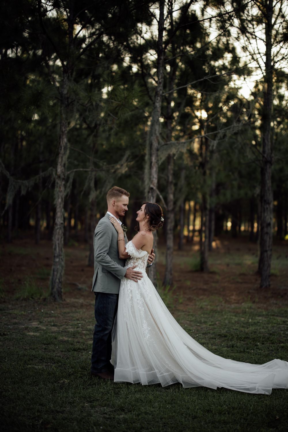 A wedding couple shares an intimate moment at sunset among tall pine trees in a forest setting.