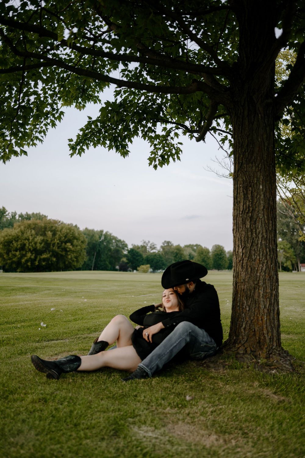 A couple relaxes together under a shady tree on green grass in a serene park setting.
