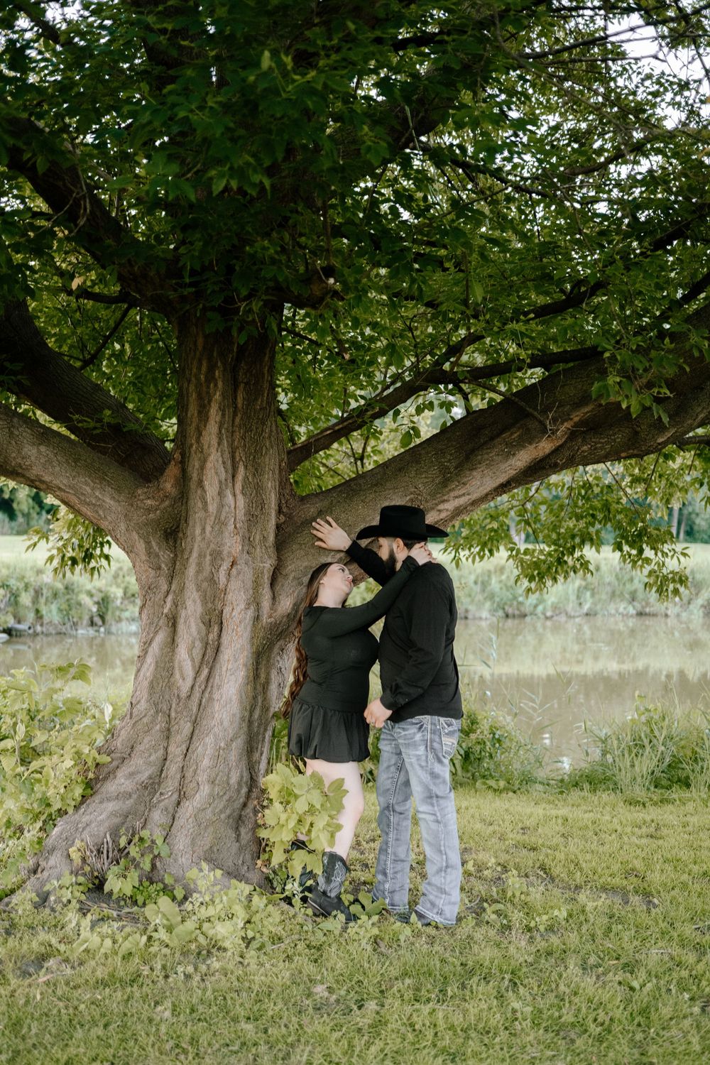 A couple in casual attire explores a large old tree in a grassy field on an overcast day.