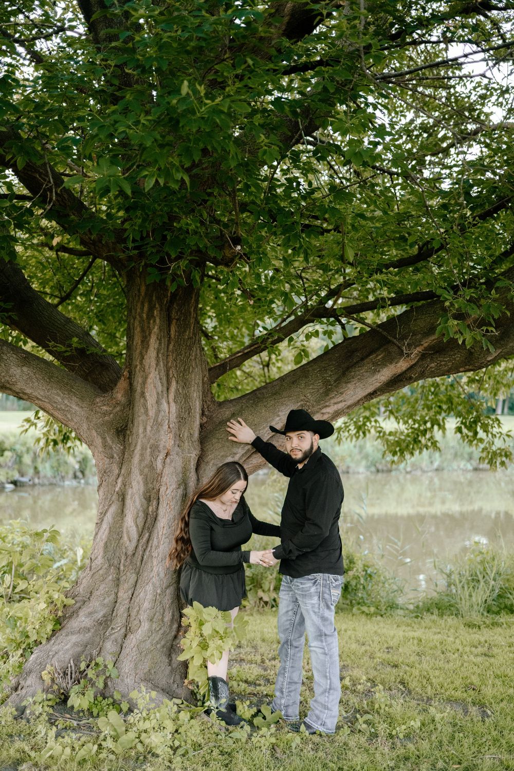 A couple shares an intimate moment beneath a large tree with sprawling branches in a serene outdoor setting.