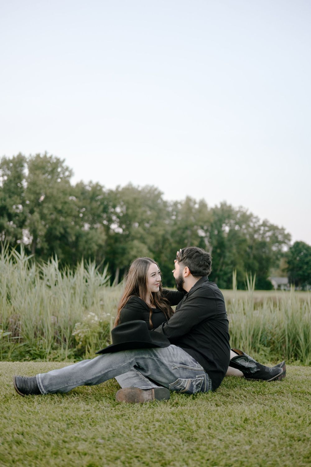 Casual couple relaxing on grass with decorative pampas grass and trees in the background.