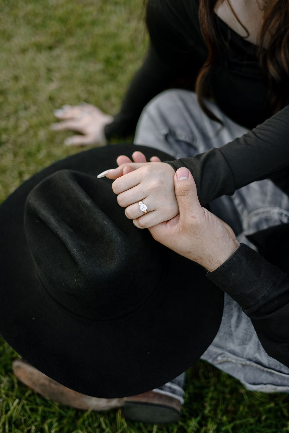 Close-up of intertwined hands with engagement ring resting on black fabric against grass.