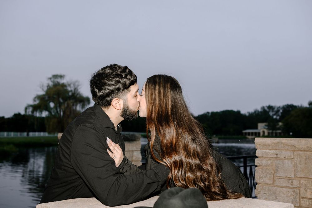 Two people share a tender moment against a stone wall with water and trees in the background.
