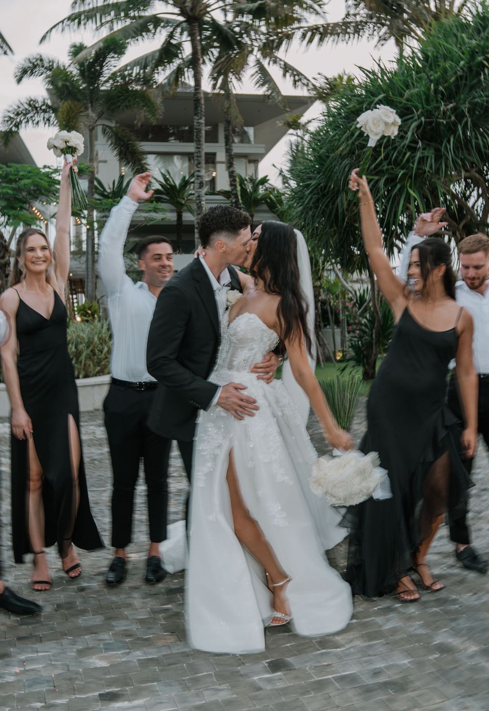 A wedding celebration with bridesmaids in black dresses and groomsmen celebrating under palm trees at a tropical venue.