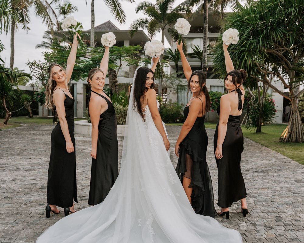 Wedding bridal party wearing black dresses and white gown walk together on stone path near palm trees.