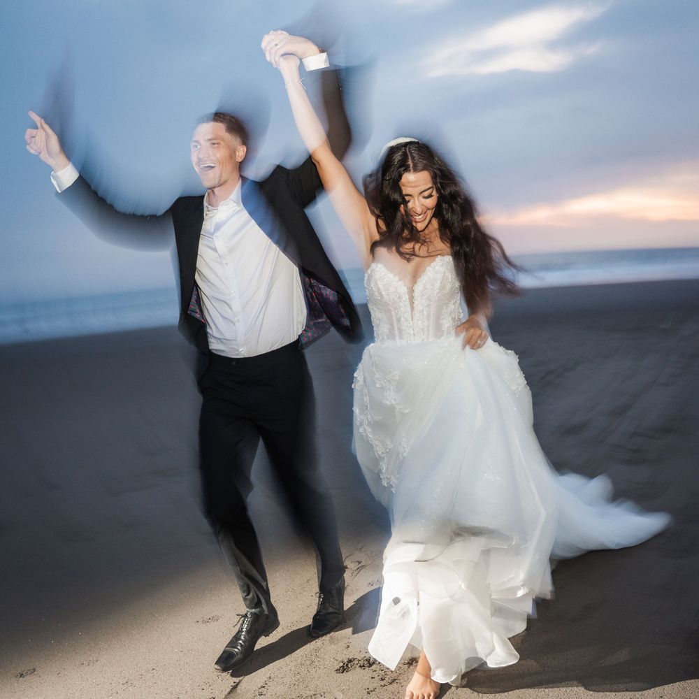 Couple dancing joyfully on a beach at sunset, twirling with the bride's white dress flowing in the breeze.