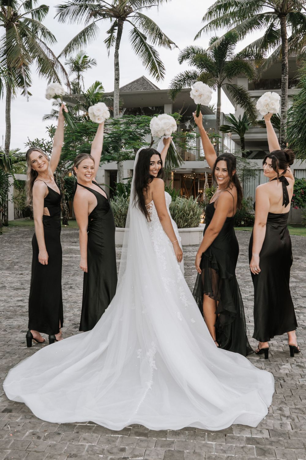 A bridal party in black dresses and white gown celebrates on a tropical beach with palm trees in the background.