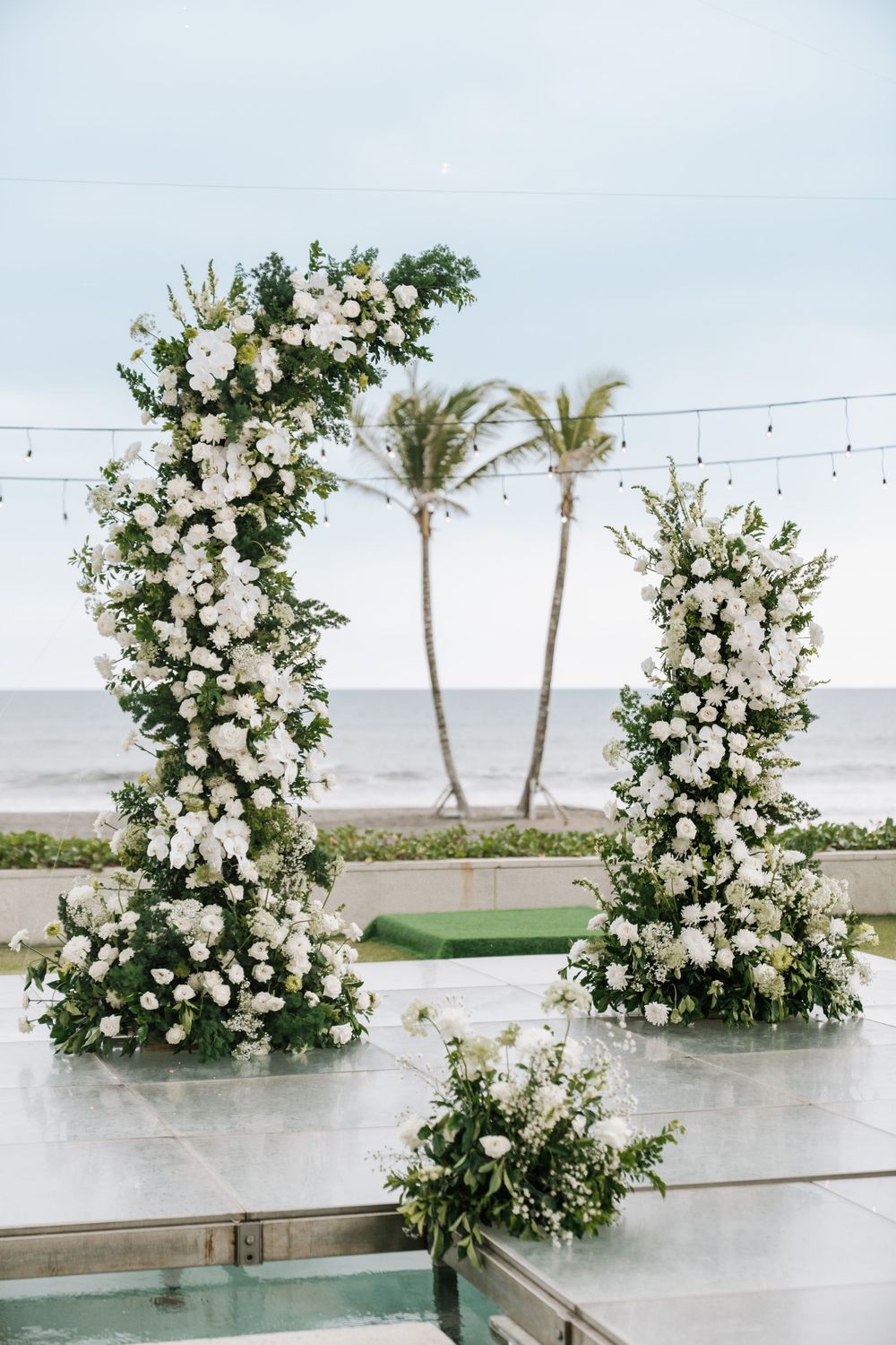 Elegant white floral arch arrangements frame a beachside wedding ceremony setup with palm trees and ocean views in the background.