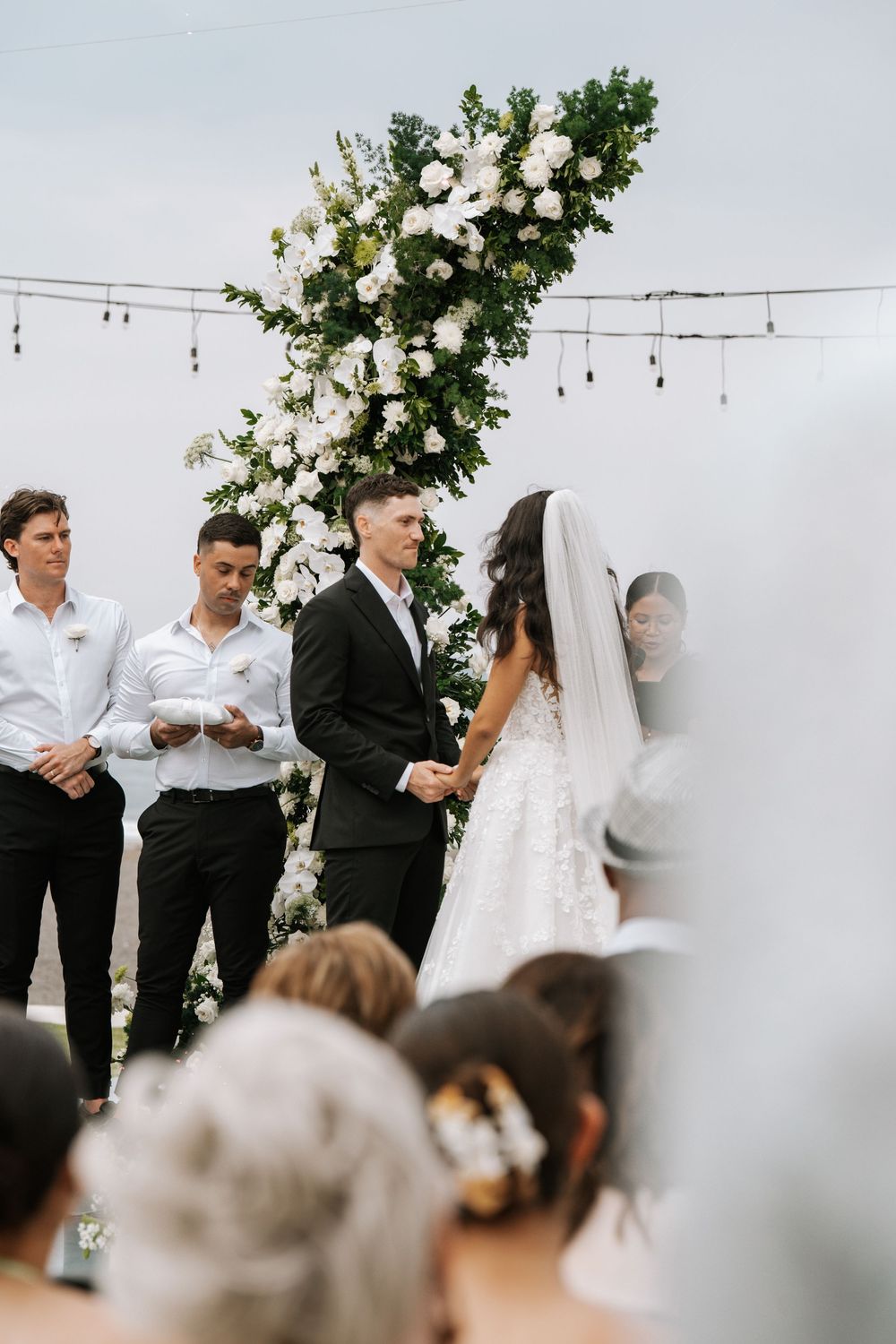 Wedding guests in formal black and white attire mingle during outdoor cocktail hour with white decorations and floral accents.