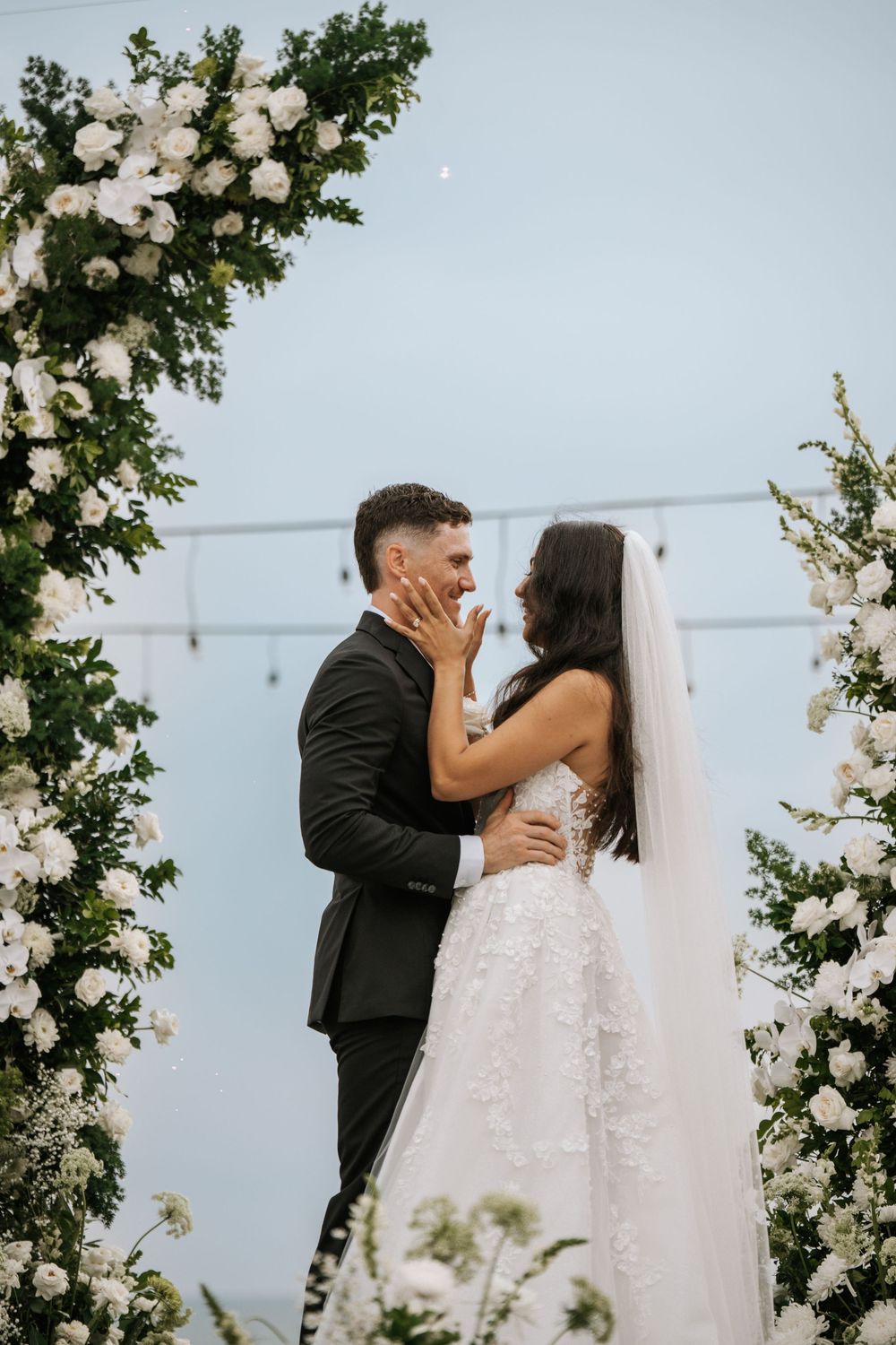 A bride and groom share their first kiss under a beautiful white floral wedding arch during their outdoor ceremony.