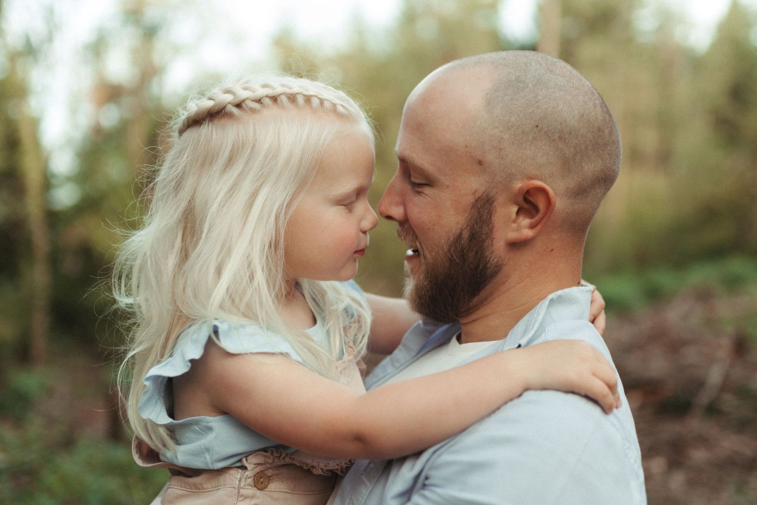 A toddler in a light-colored outfit laughs joyfully while holding onto an adult's hands outdoors in natural lighting.