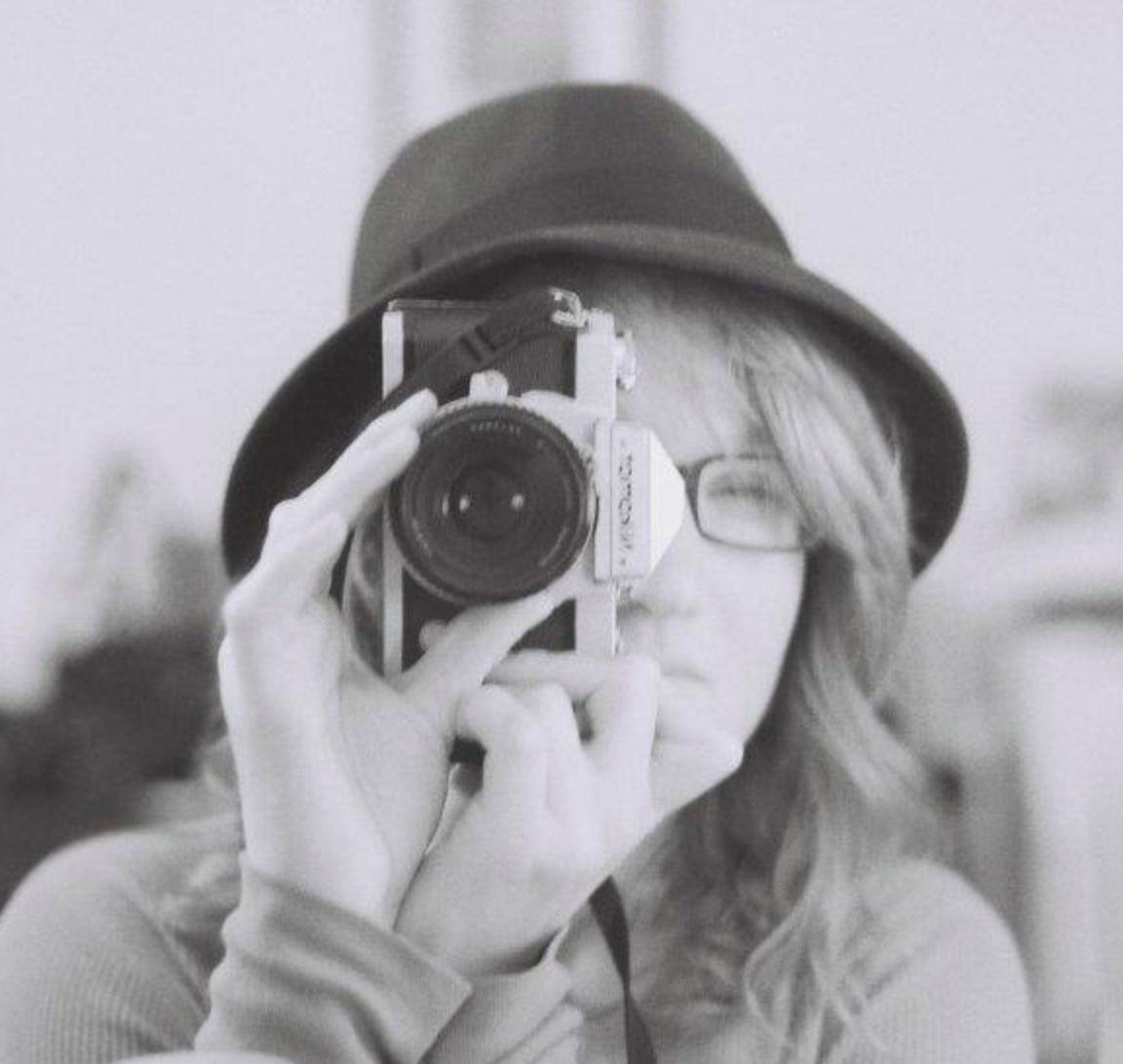 Black and white photo of a photographer holding a camera with a large lens wearing a dark hat.