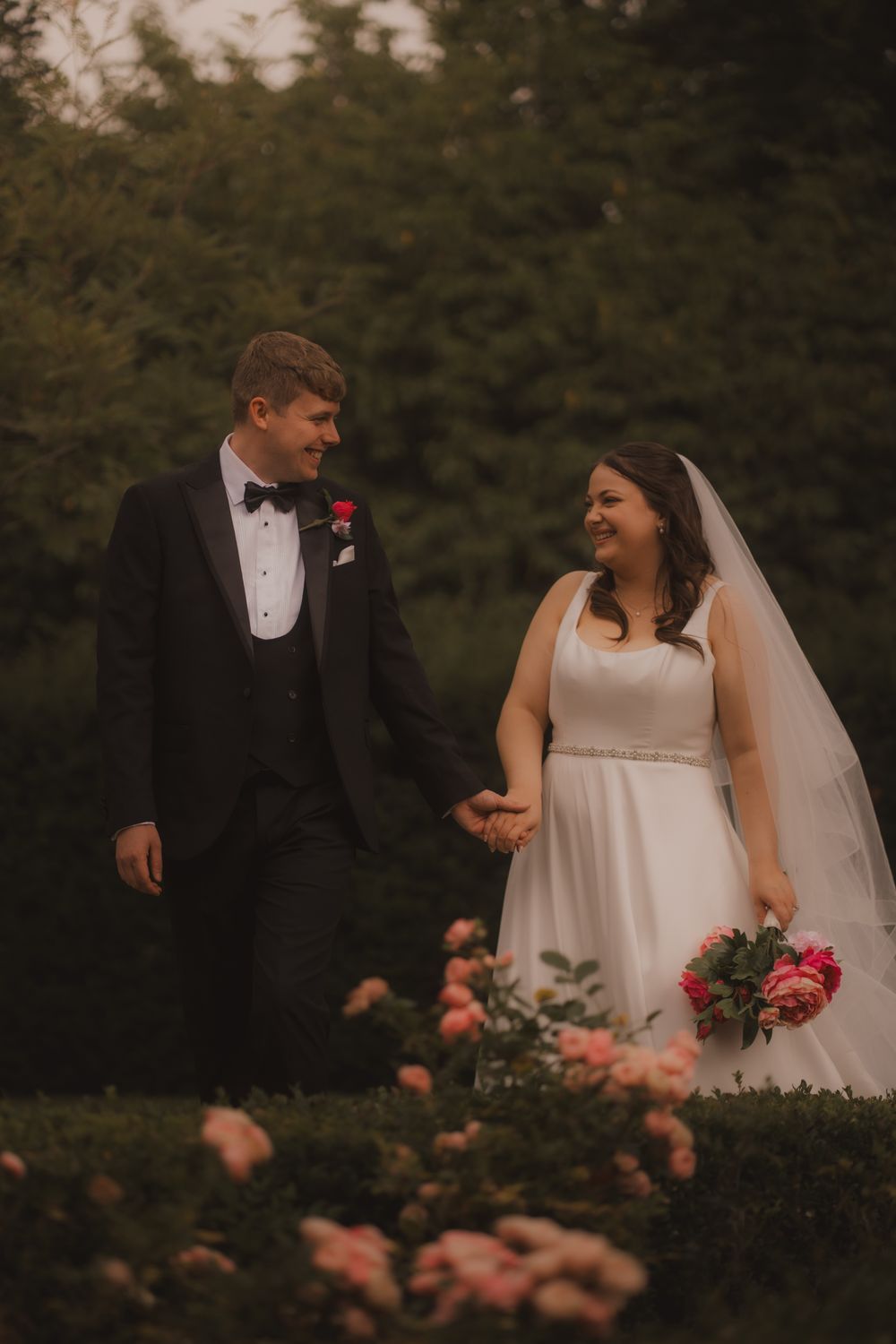 A bride and groom share intimate moments in a garden setting with blooming pink roses at sunset.