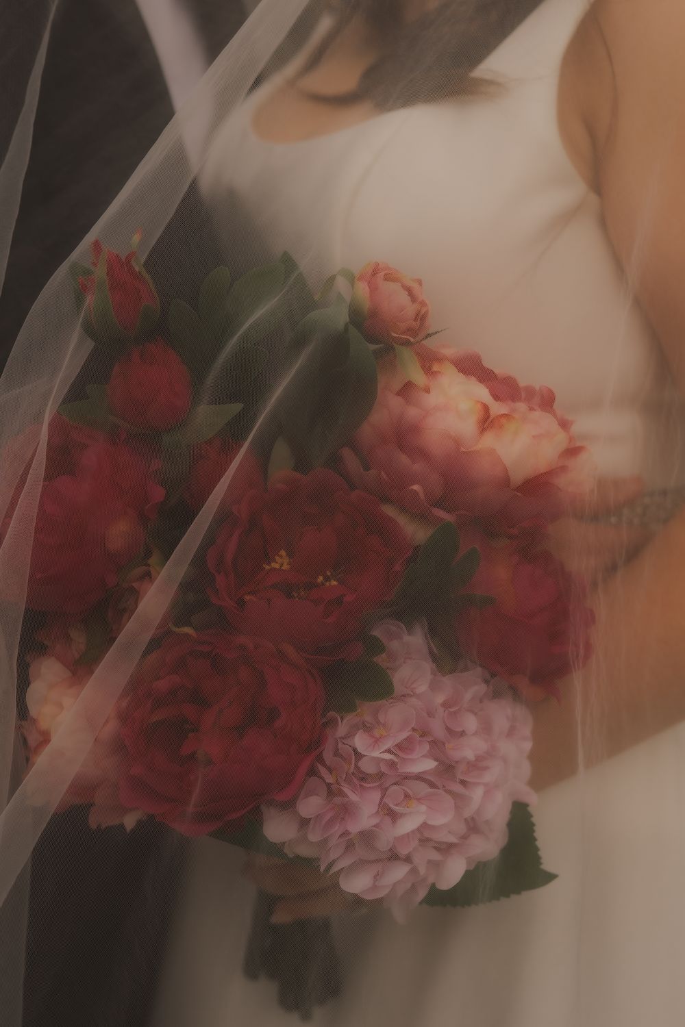 A dreamy, soft-focus bouquet of red and pink roses and peonies held against a white dress with sheer fabric overlay.