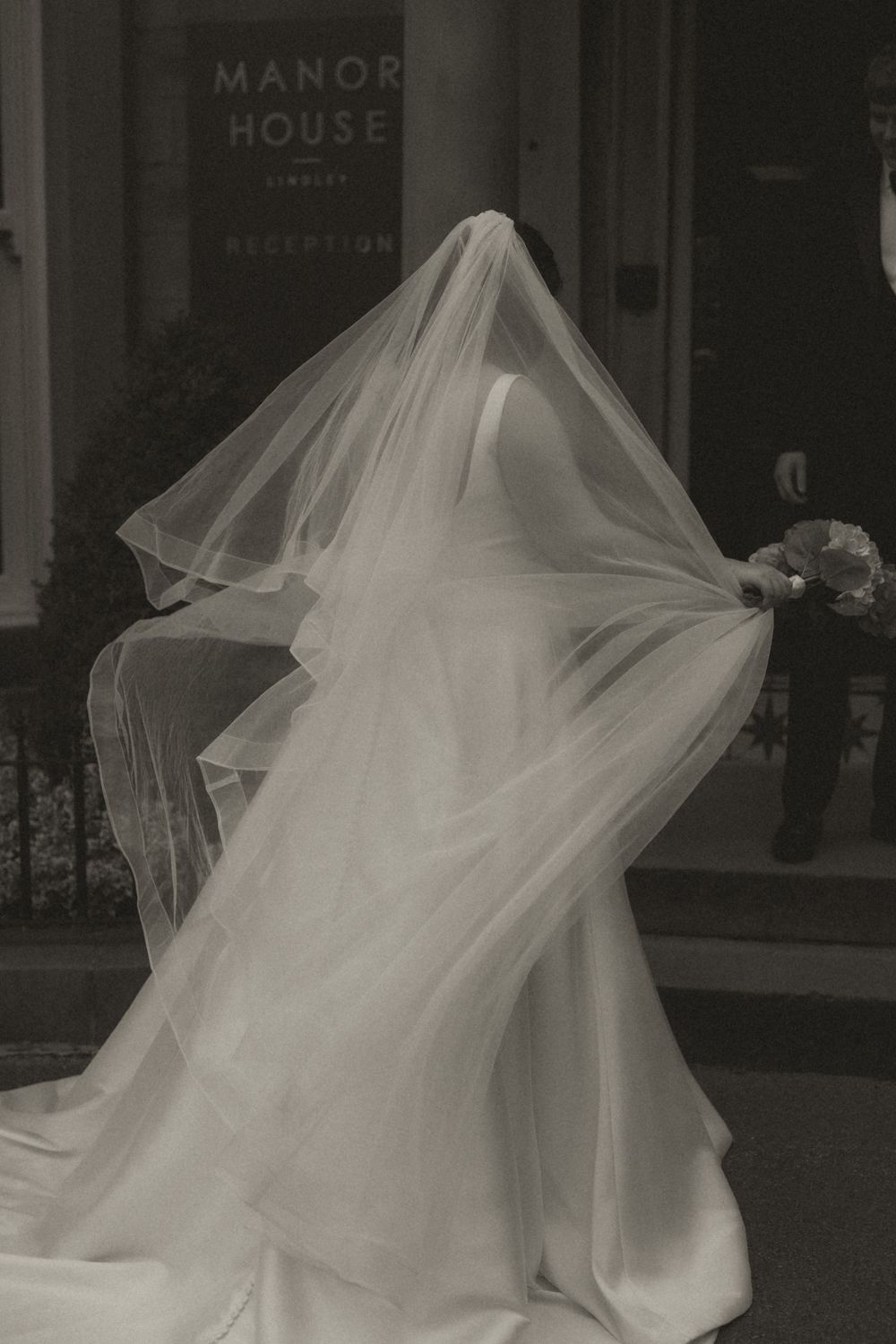 A flowing white wedding dress with veil billows dramatically in motion against a storefront backdrop.