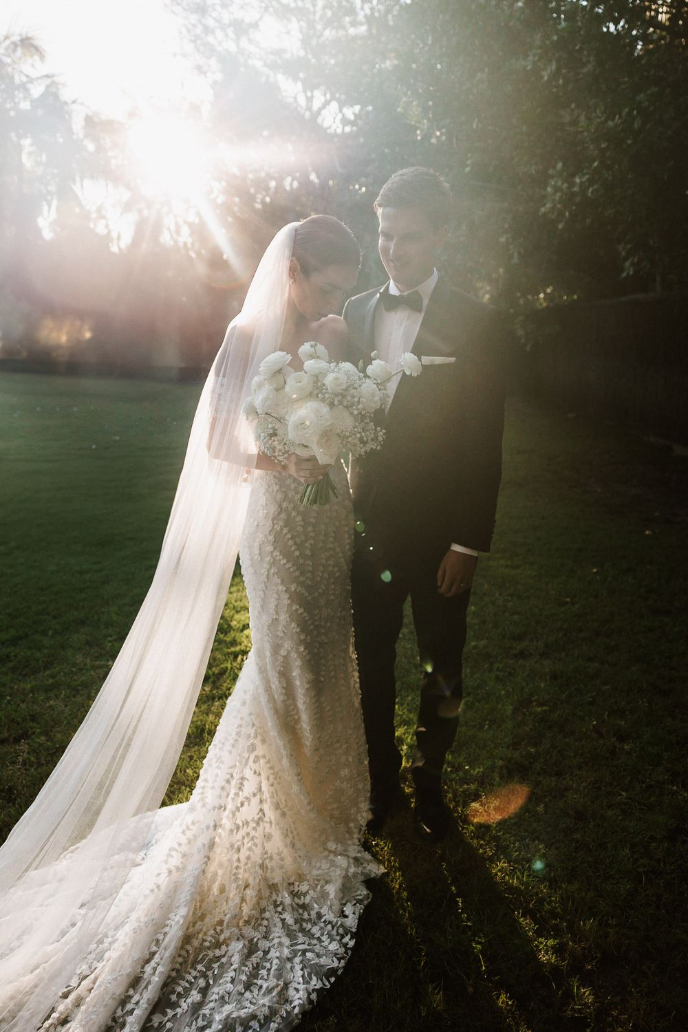 Ethereal sunlit portrait of bride and groom outdoors with flowing lace wedding dress and veil in golden hour light.