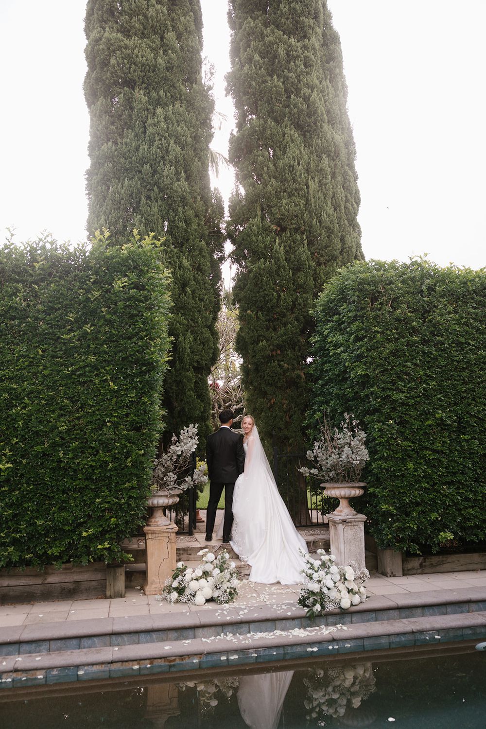 Wedding couple standing by tall cypress trees and hedges near a reflecting pool with floral decorations.