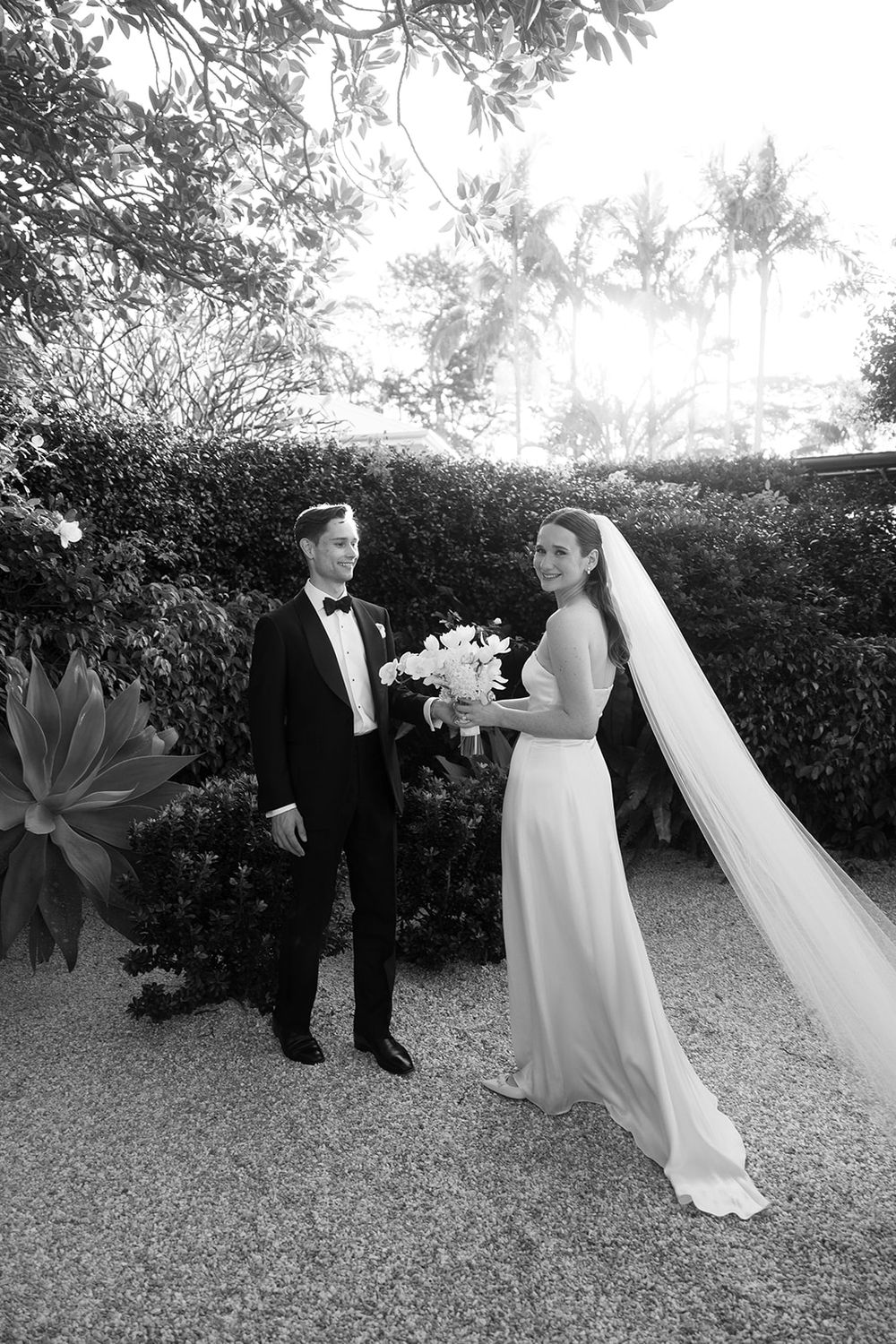 Black and white photo of a bride in white gown with veil and groom in tuxedo posing outdoors near lush hedges.