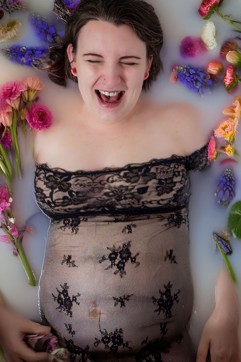 A woman lays in a milk bath surrounded by vibrant flowers with an expression of joy and laughter on her face.