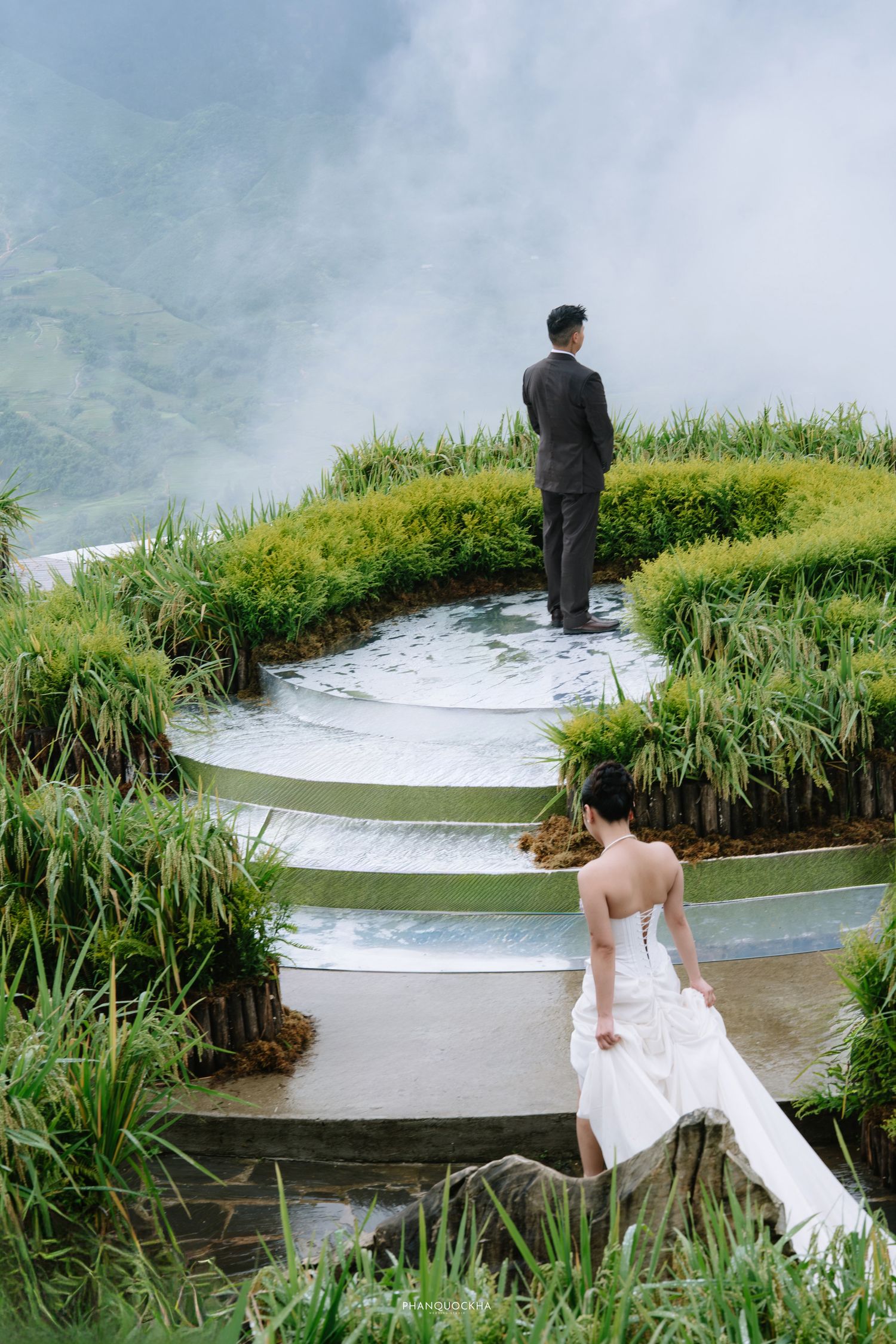 Bride in white gown walks toward groom on terraced rice field path with misty mountain backdrop.