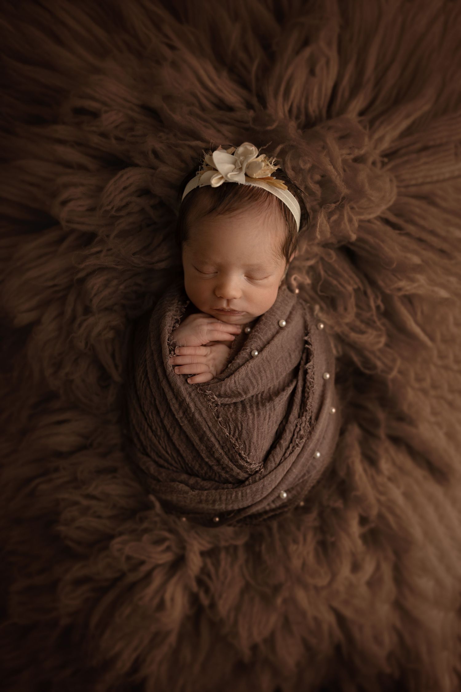 Artistic newborn photography with baby wrapped in brown fabric on fluffy textured background.