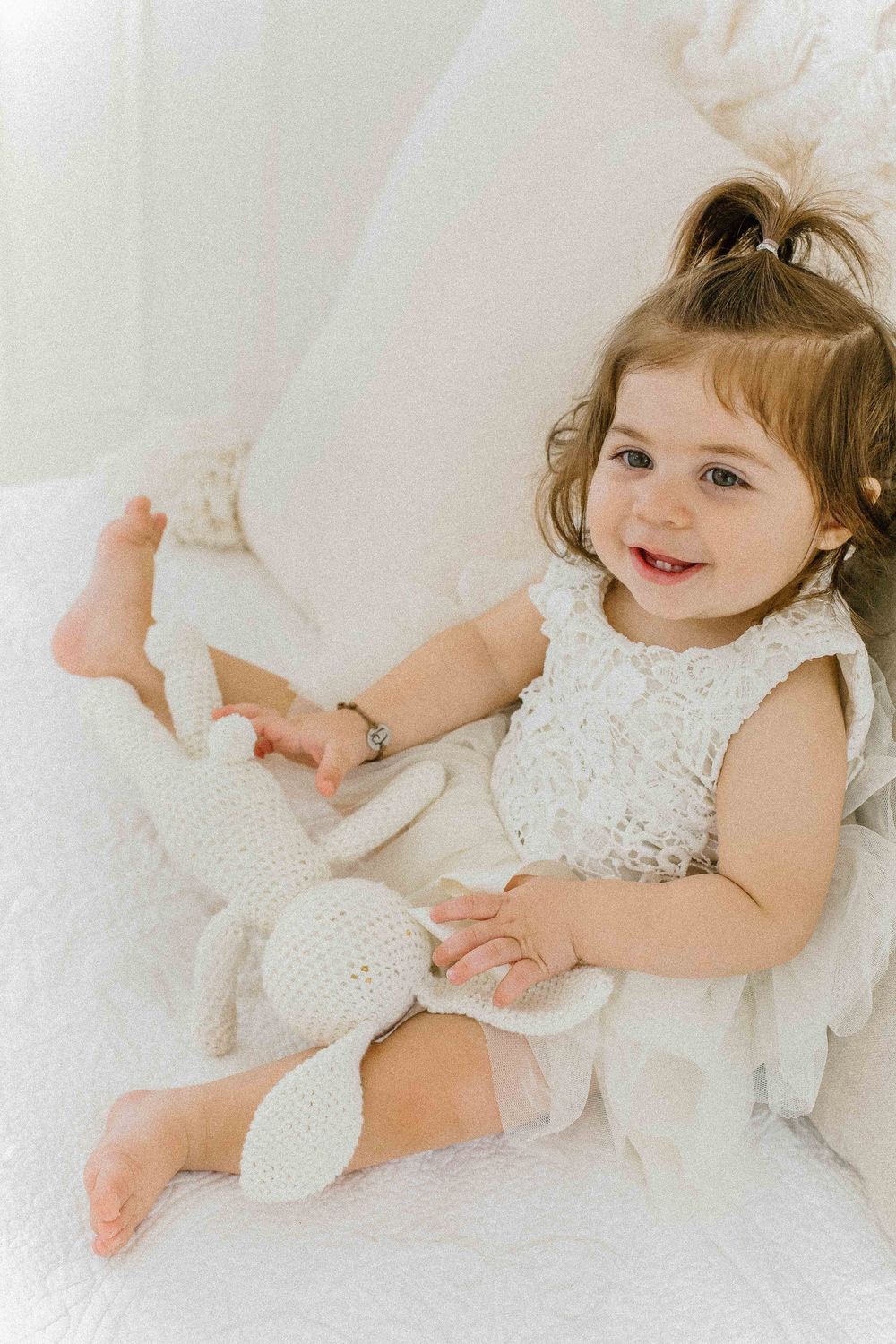 Young child in white lace dress lying on white bedding during portrait session. Create timeless memories with our maternity, newborn.