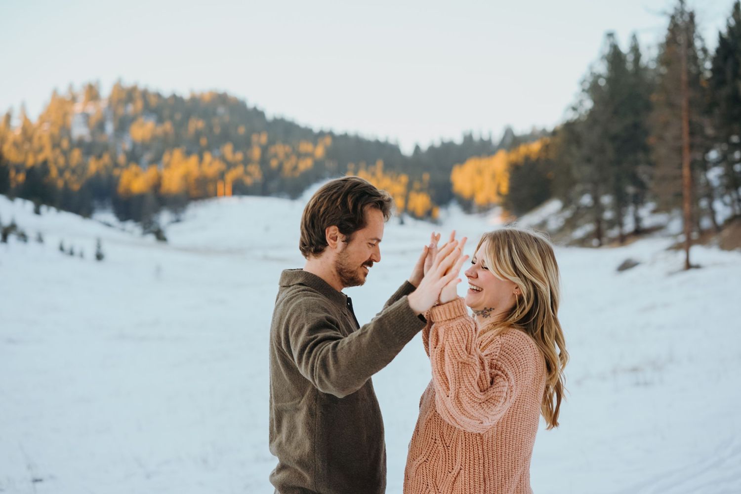 A playful couple shares a joyous moment together in the snow with autumn trees on a mountain slope in the background.